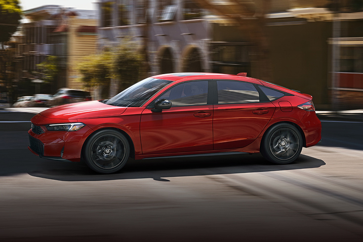 2025 Honda Civic Hatchback Sport Hybrid in Rallye Red profile driving on a city street (Compact Hatchback).